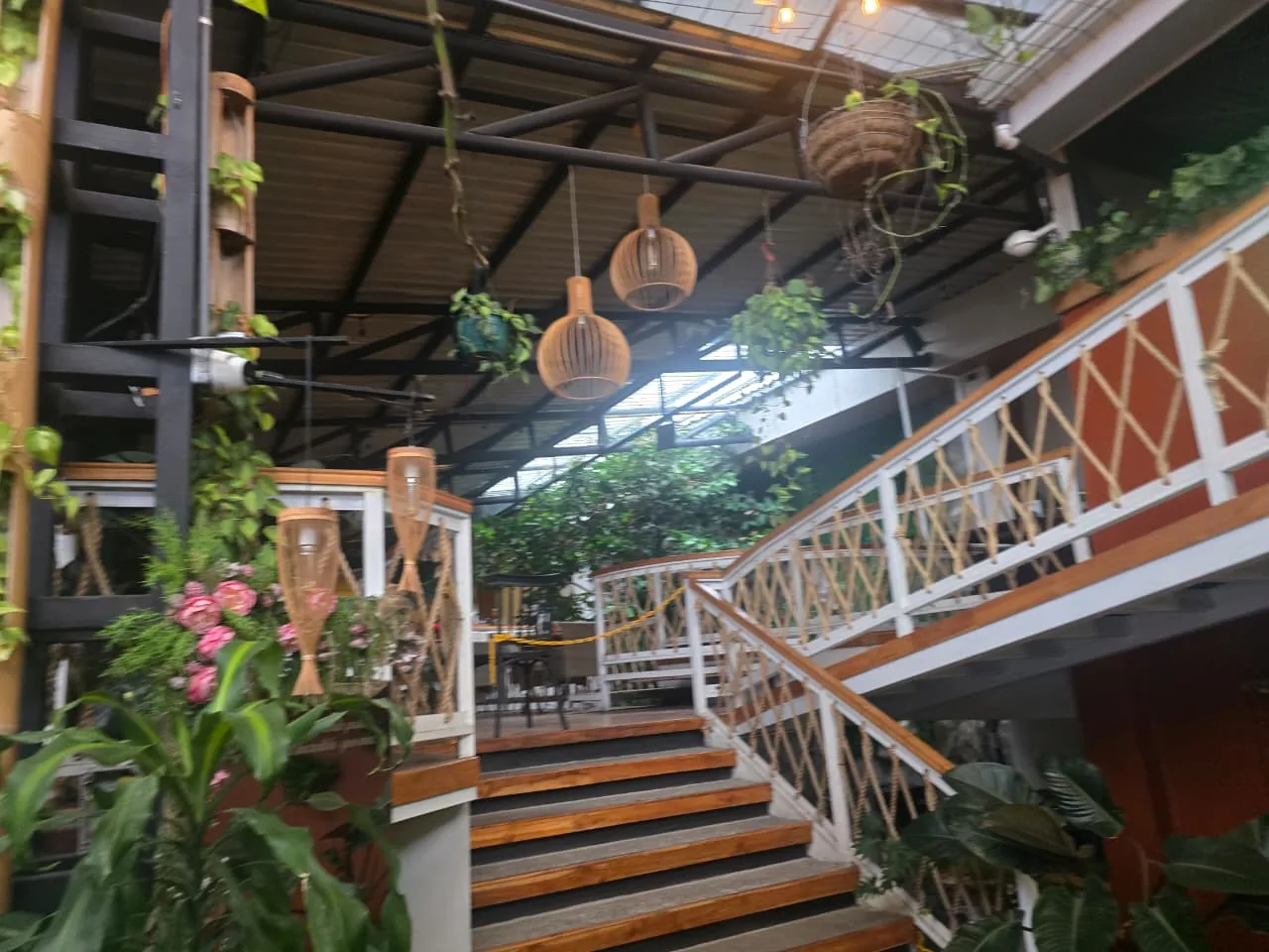 Interior del mercado: gradas de madera que suben a una tarima con mesas, barandas blancas tejidas con cuerda, lámparas colgantes de mimbre y plantas colgando de la estructura metálica del techo.
